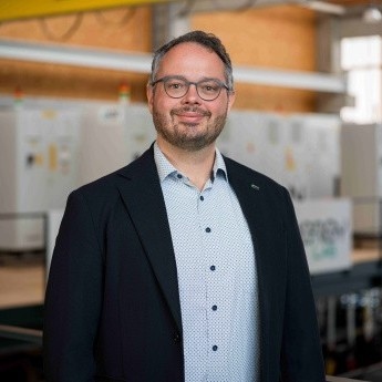 Porträt eines Mannes mit Brille und Sakko vor industrieller Anlage.
